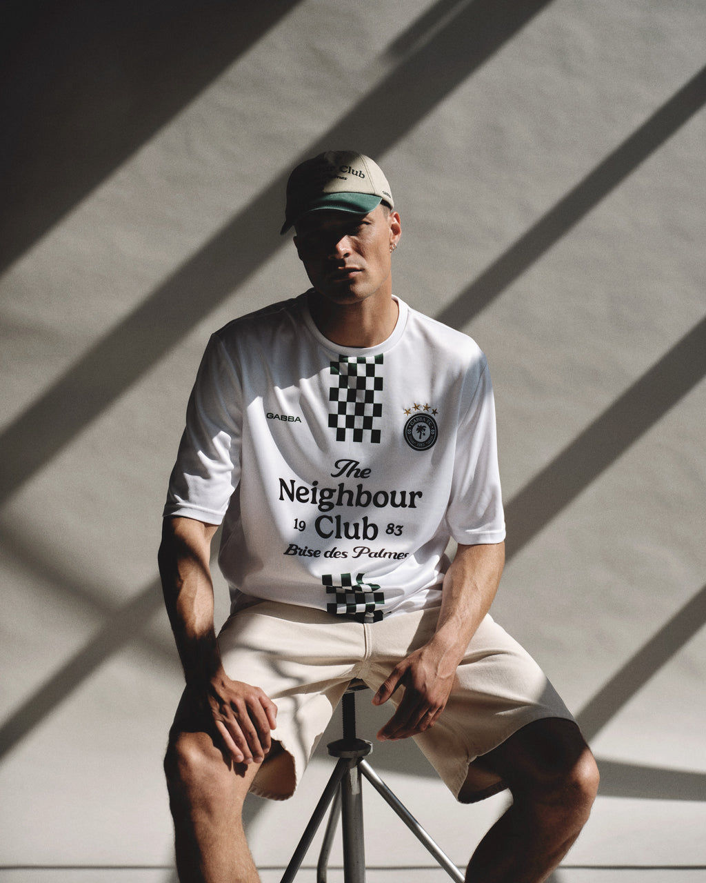 A man in a beige cap, beige shorts, and a Gabba GABnigel Fut Boxy Pes SS T-shirt in white sits on a stool with hands on his knees; sunlight casts diagonal shadows across the plain background.