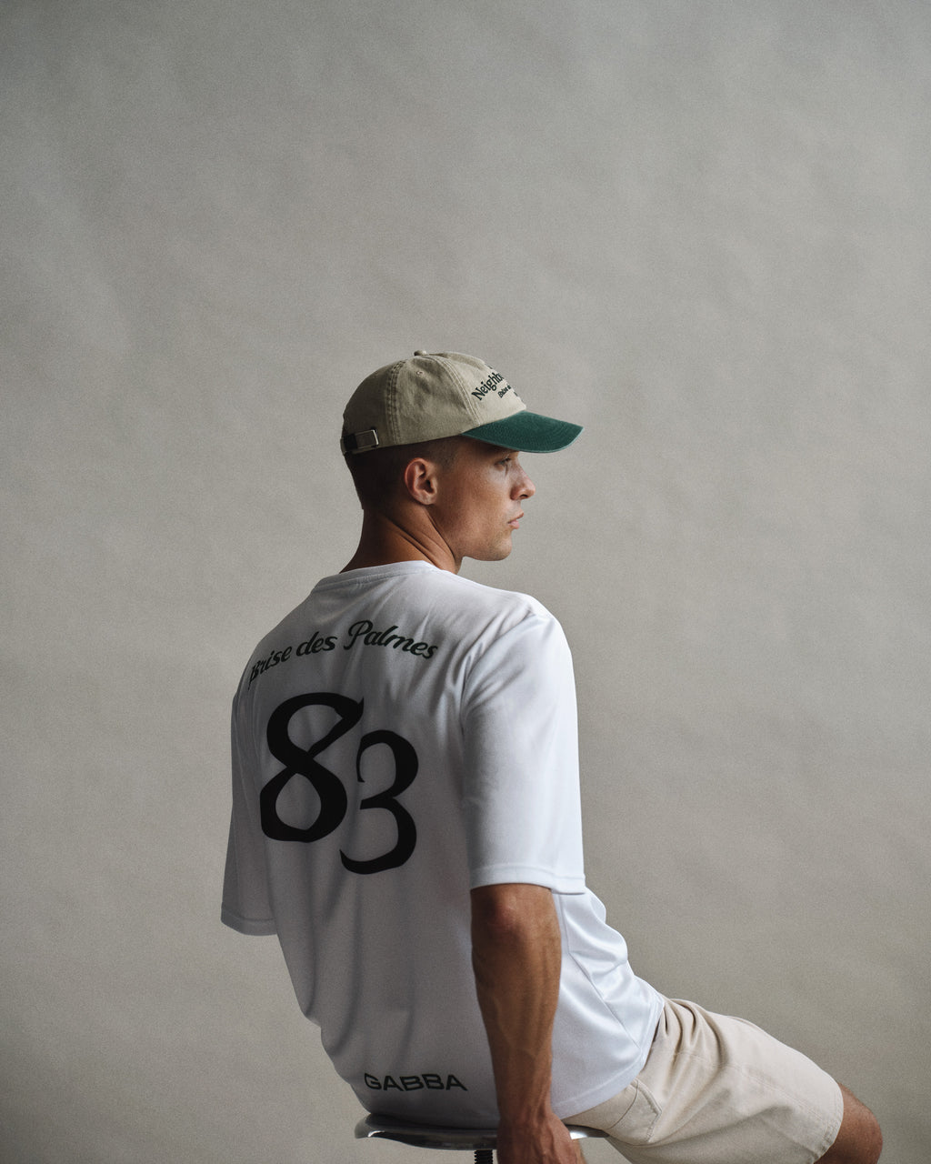 A man in a beige cap and light shorts sits sideways on a stool against a plain background, wearing the Gabba GABnigel Fut Boxy Pes SS T-shirt in white, which features “83” and French text on the back.
