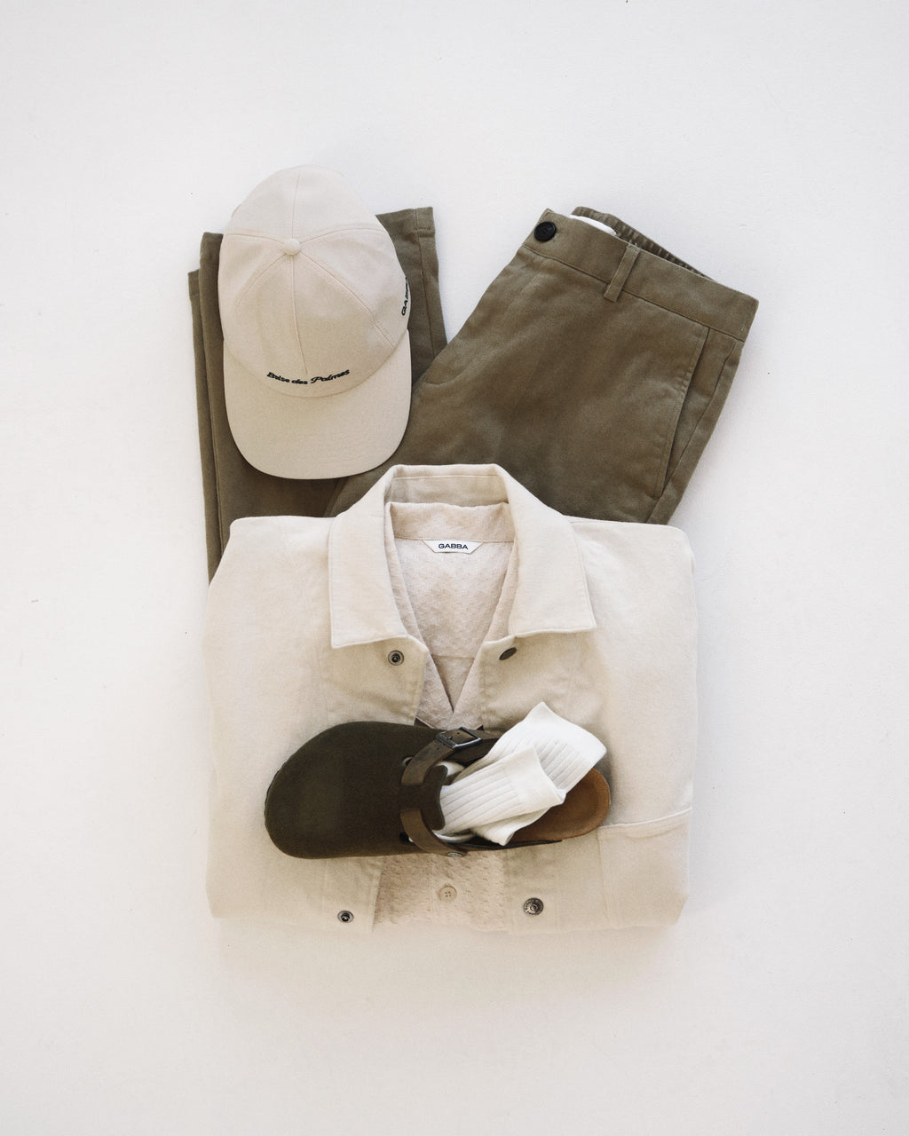 A light beige shirt, Gabba GABdenver Elastic Twill Pant in green, a beige baseball cap, white socks, and dark brown sandals are arranged on a white surface.