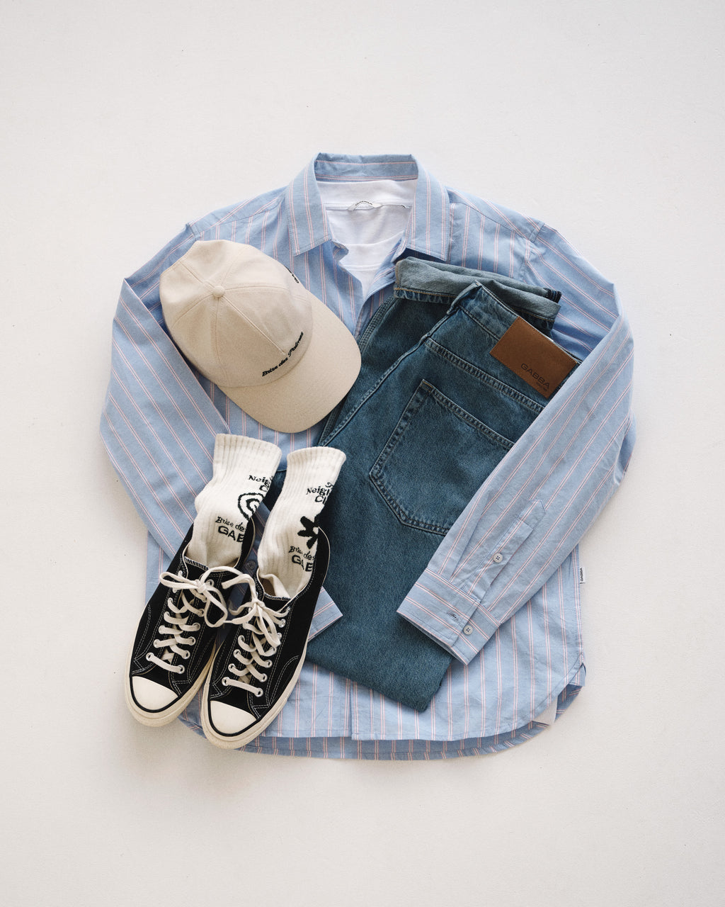 A laid-out outfit with a blue and white striped button-up shirt, Gabba GABnoah Brook Jeans in denim wash, a beige baseball cap, white socks, and black Converse sneakers, all arranged neatly on a white background.