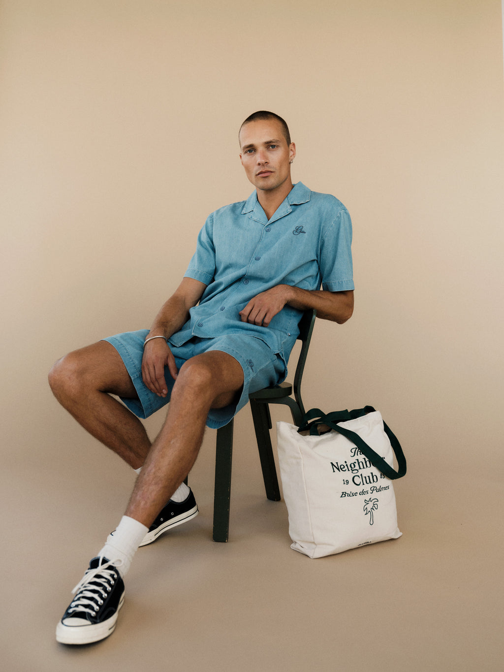 A man wearing the Gabba GABtimo Resort Denim SS Shirt in light denim blue sits on a green chair against a beige background. He pairs it with matching shorts, black sneakers, white socks, and has a white tote bag with green handles beside him.