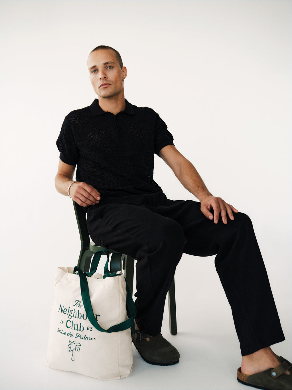 A person with short hair in a black Gabba GABbenson Fiwi SS polo and dark shoes sits on a green chair holding a tote bag reading 