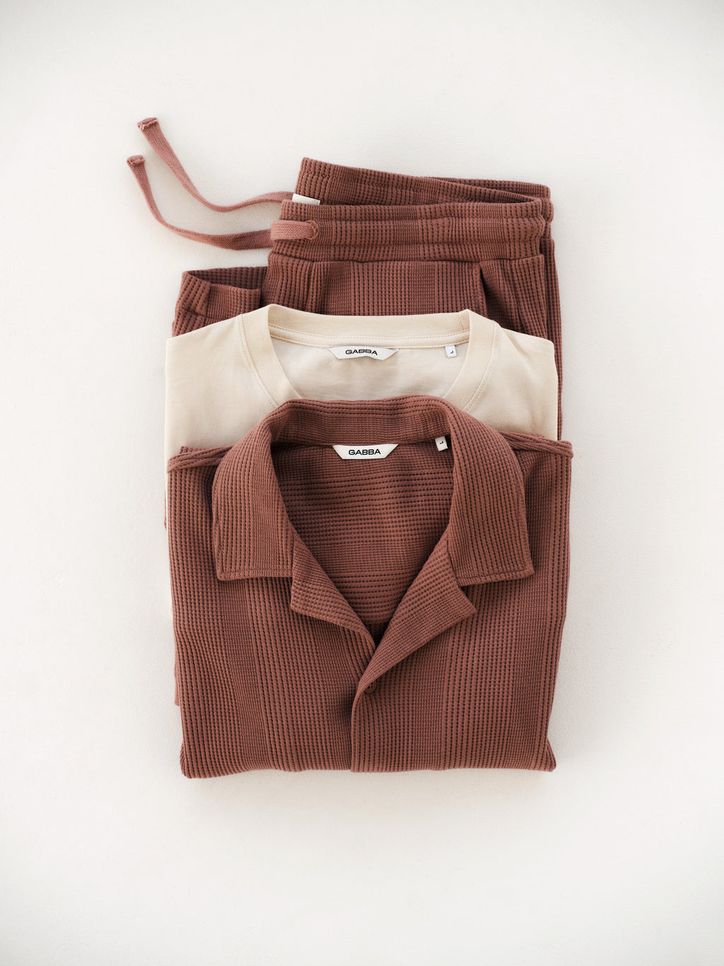 Neatly folded on a white surface: Gabba's GABtommi Wup Shorts in Wood Pink, paired with a beige t-shirt and matching button-up shirt.