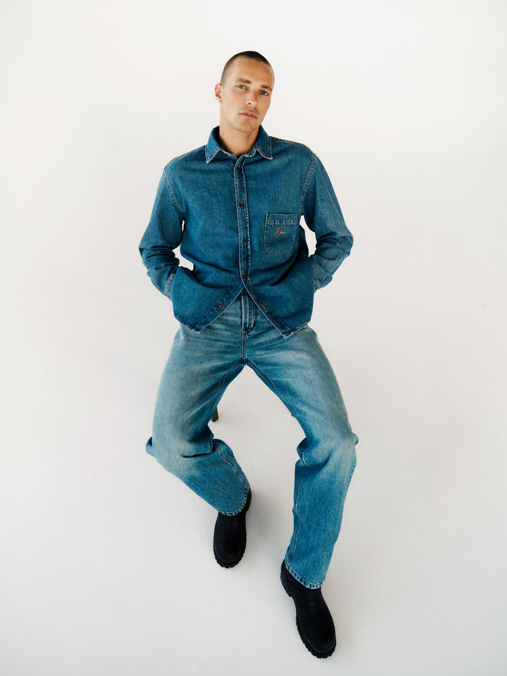 A person with short hair wears a blue denim twill jacket and Gabba GABmax Elvis Jeans in Mid Blue Denim, sitting on a white surface with hands in pockets, looking to the side. Black boots complete the look.