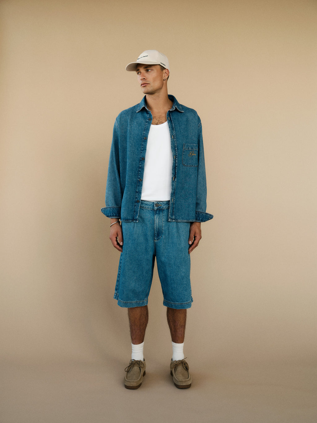 A man stands against a beige background in relaxed style: light beige cap, open denim shirt over white T-shirt, Gabba GABmoro PasBrook Shorts – Denim wash, with white socks and beige shoes. He faces sideways, hands by his sides.
