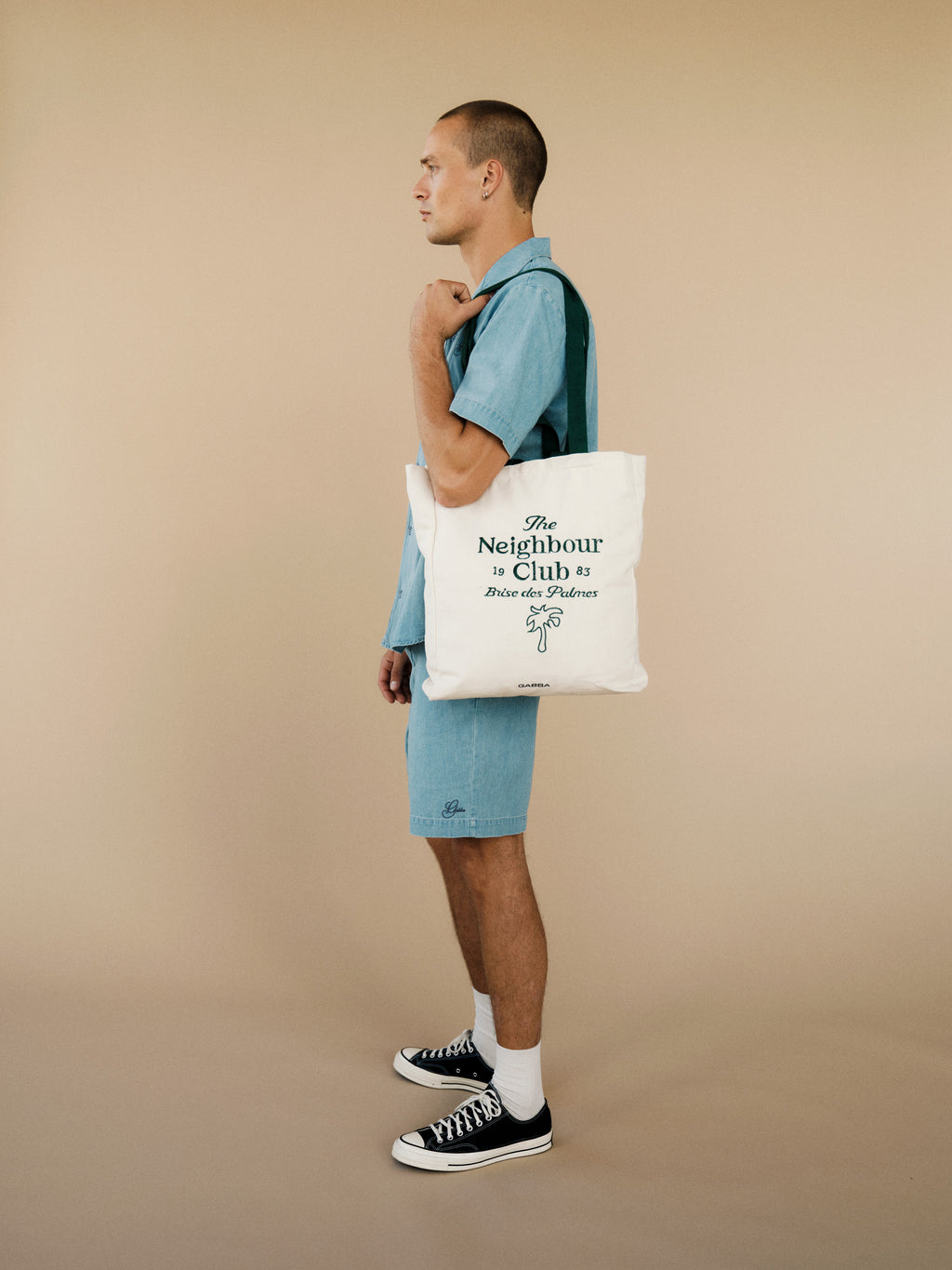 A person wears the Gabba GABtimo Resort Denim SS Shirt in Lt. Denim Blue, paired with shorts, black sneakers, and white socks. They stand sideways against a beige background holding a white tote bag with black text.