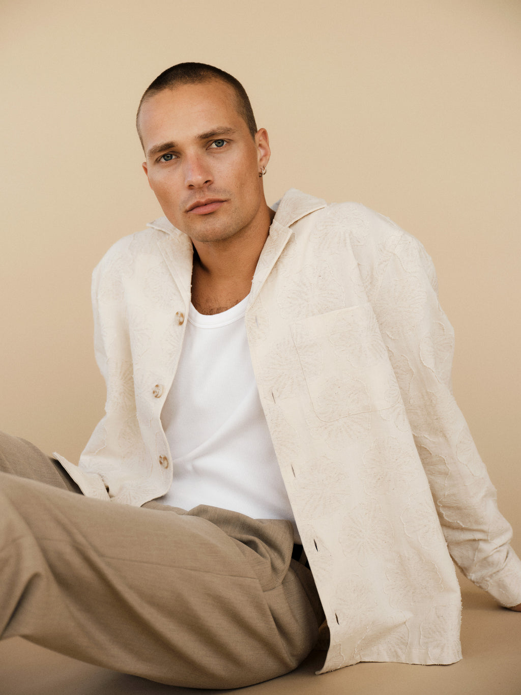 A person with a buzz cut and neutral expression wears the Gabba GABaustin Reso Raw LS Overshirt in Lt. Sand over a white top and beige pants, seated against a beige background.