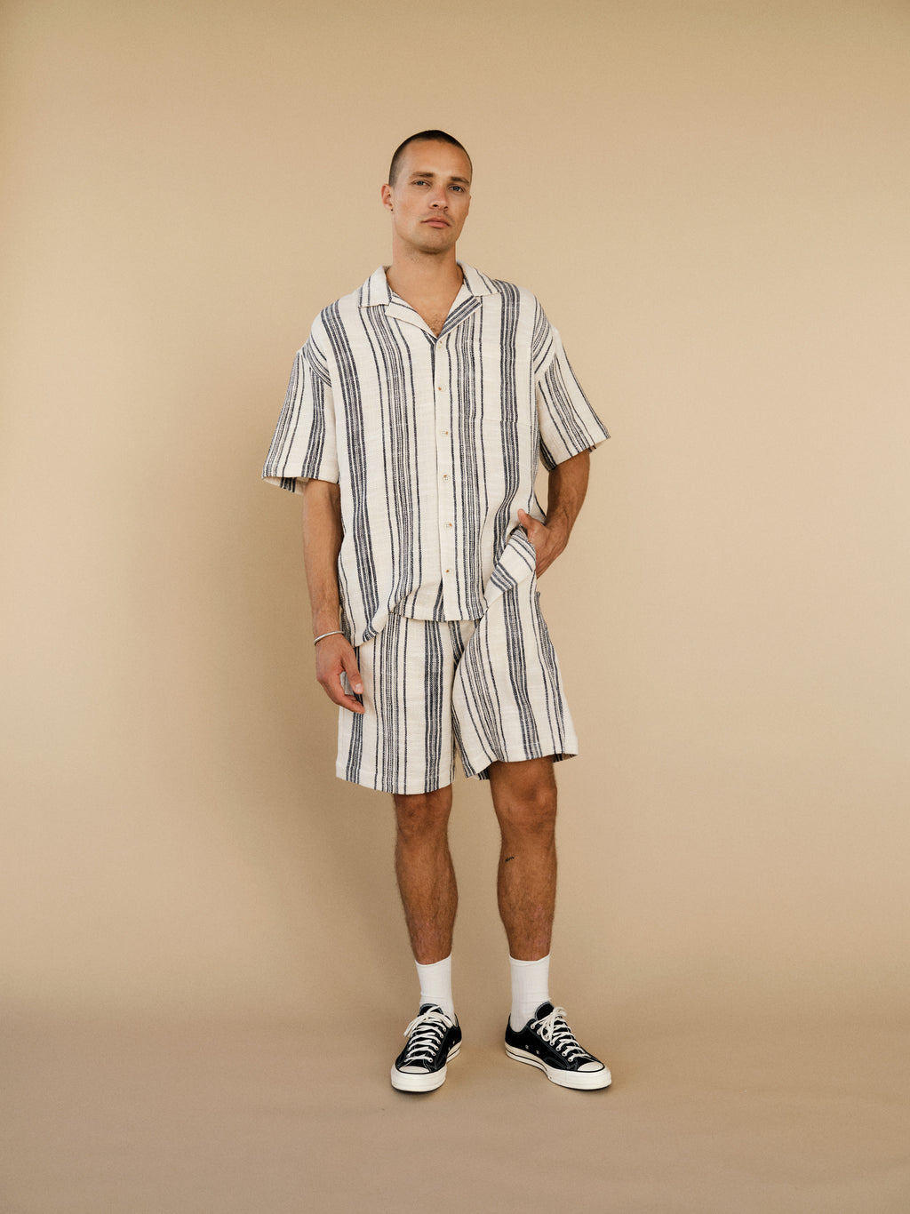 A man stands before a beige background wearing Gabba’s GABtommi Riga Shorts in navy stripe with an elastic waistband, paired with a matching short-sleeve button-up. He has one hand in his pocket and sports black sneakers with white socks.