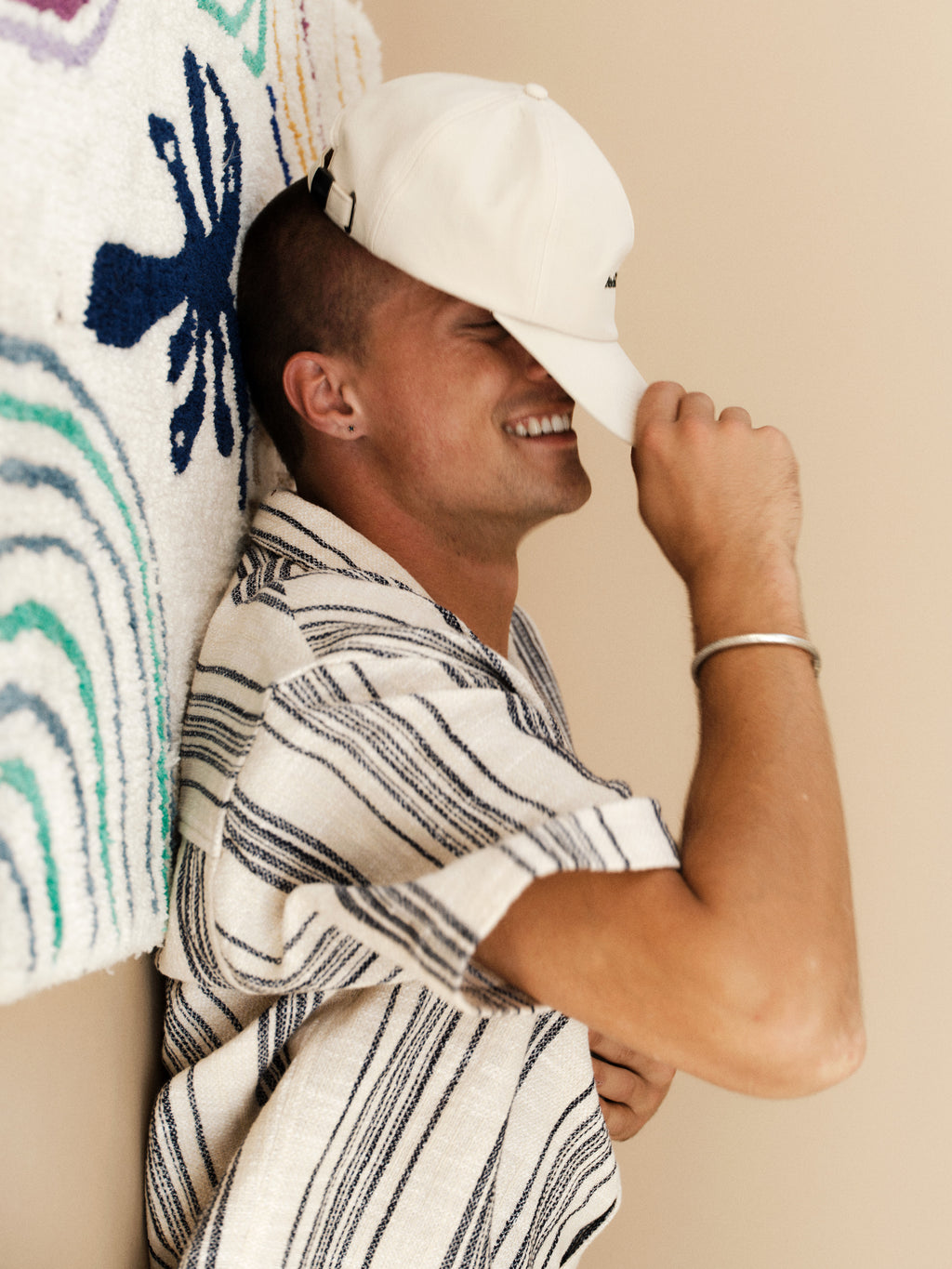 A smiling person in a striped shirt and the Gabba GABpalms Club Cap - Birch tilts the cap over their eyes while leaning against a wall adorned with textured, colorful, soft fabric.