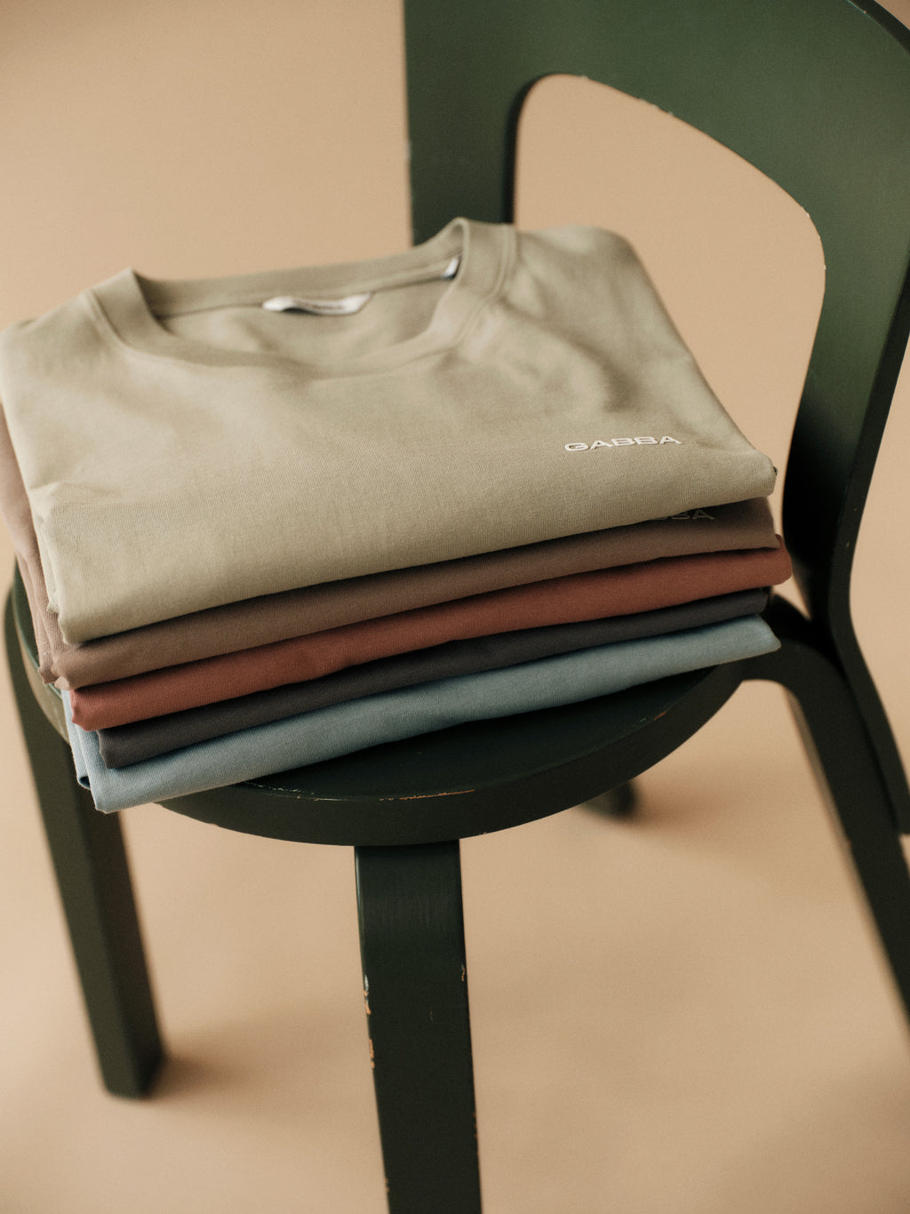 A Gabba GABdune Logo SS GOTS light green organic cotton t-shirt is neatly folded on a dark green wooden chair with a rounded seat and curved backrest, set against a beige background.