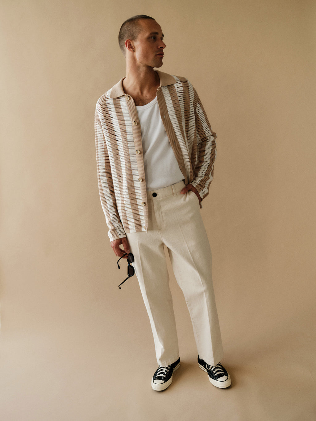 A man stands against a beige background, wearing the Gabba GABdezz Bold LS Knit in Beige Stripe over a white tank top, cream pants, and black sneakers. He holds sunglasses in one hand and looks to the side.