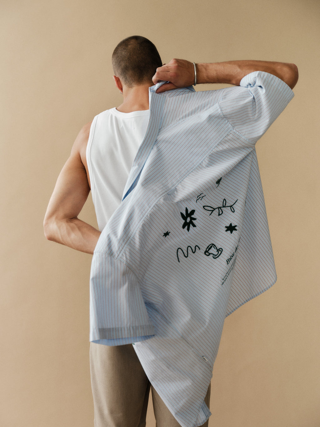 A man with short hair, seen from behind, wears a white tank top and Gabba GABjoli Pas Phin Shorts GRS in Sand Plaza while slipping on a relaxed-fit light blue pinstripe shirt with black doodles, standing against a plain beige background.