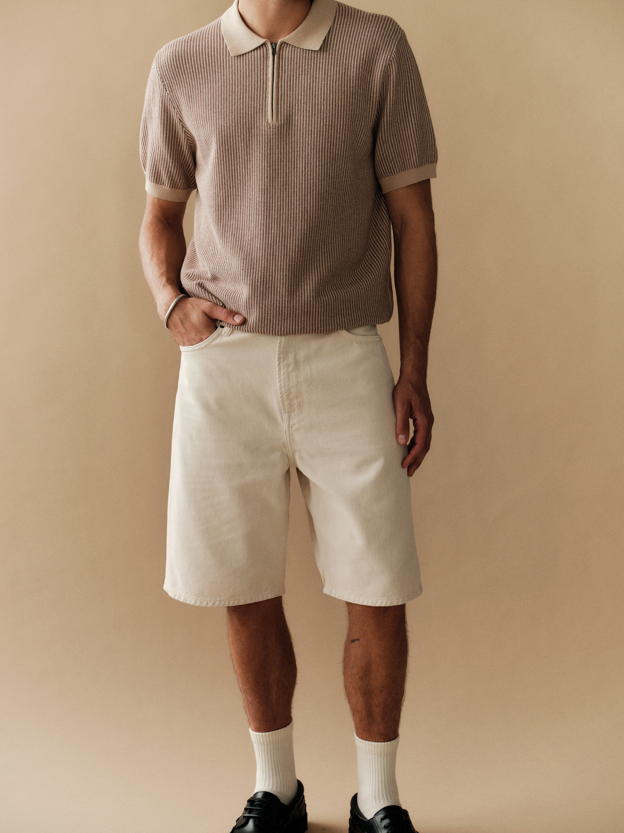 A person stands against a beige background wearing Gabba GABmox Antiq Shorts in Ecru with a taupe polo, white socks, and black shoes. One hand is in their pocket; their head is not visible in the image.