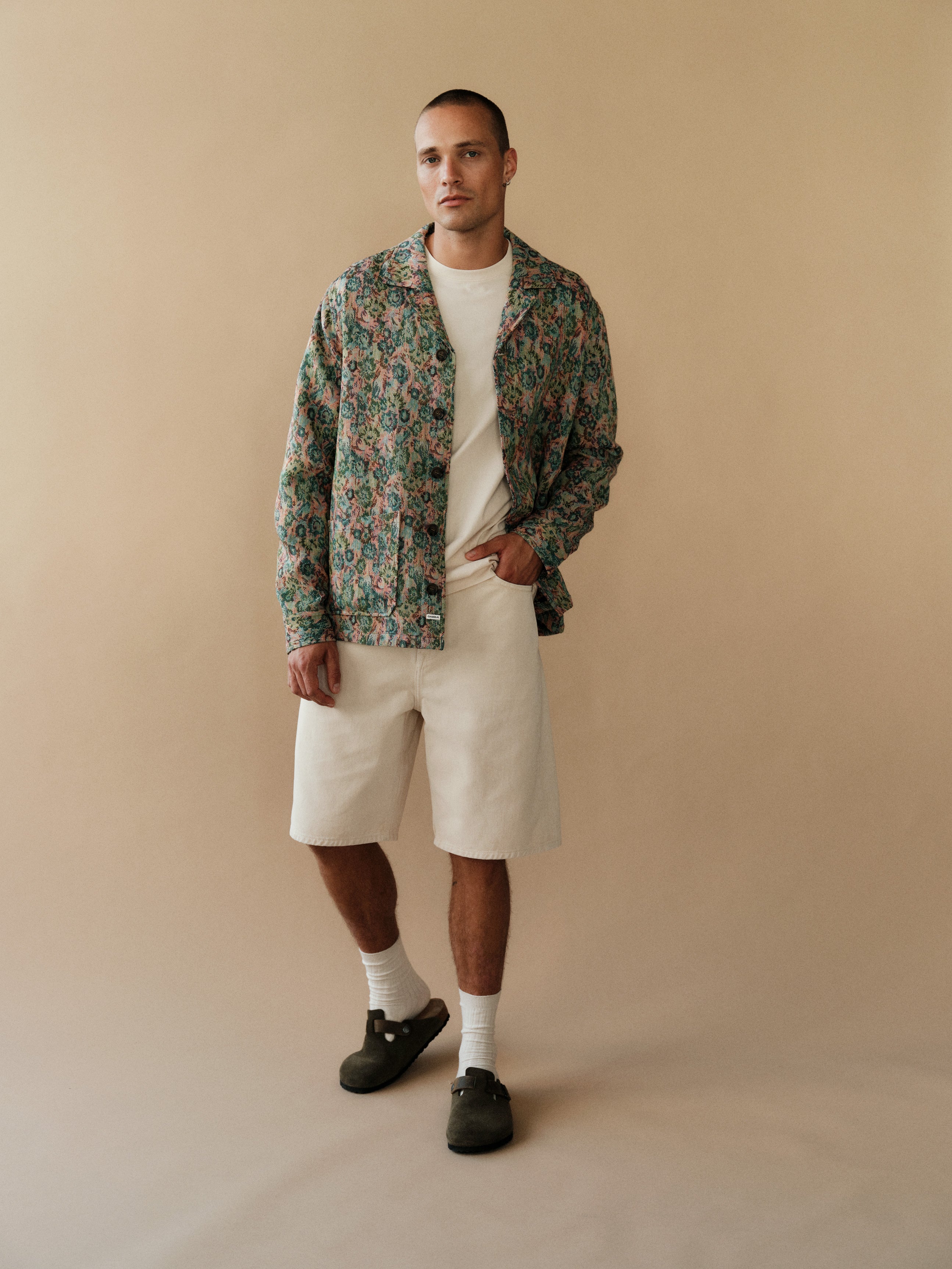 A man with short hair stands casually against a beige background, wearing the Gabba GABdune Logo SS GOTS white t-shirt, patterned jacket, off-white shorts, white socks, and dark slip-on shoes with one hand in his pocket.