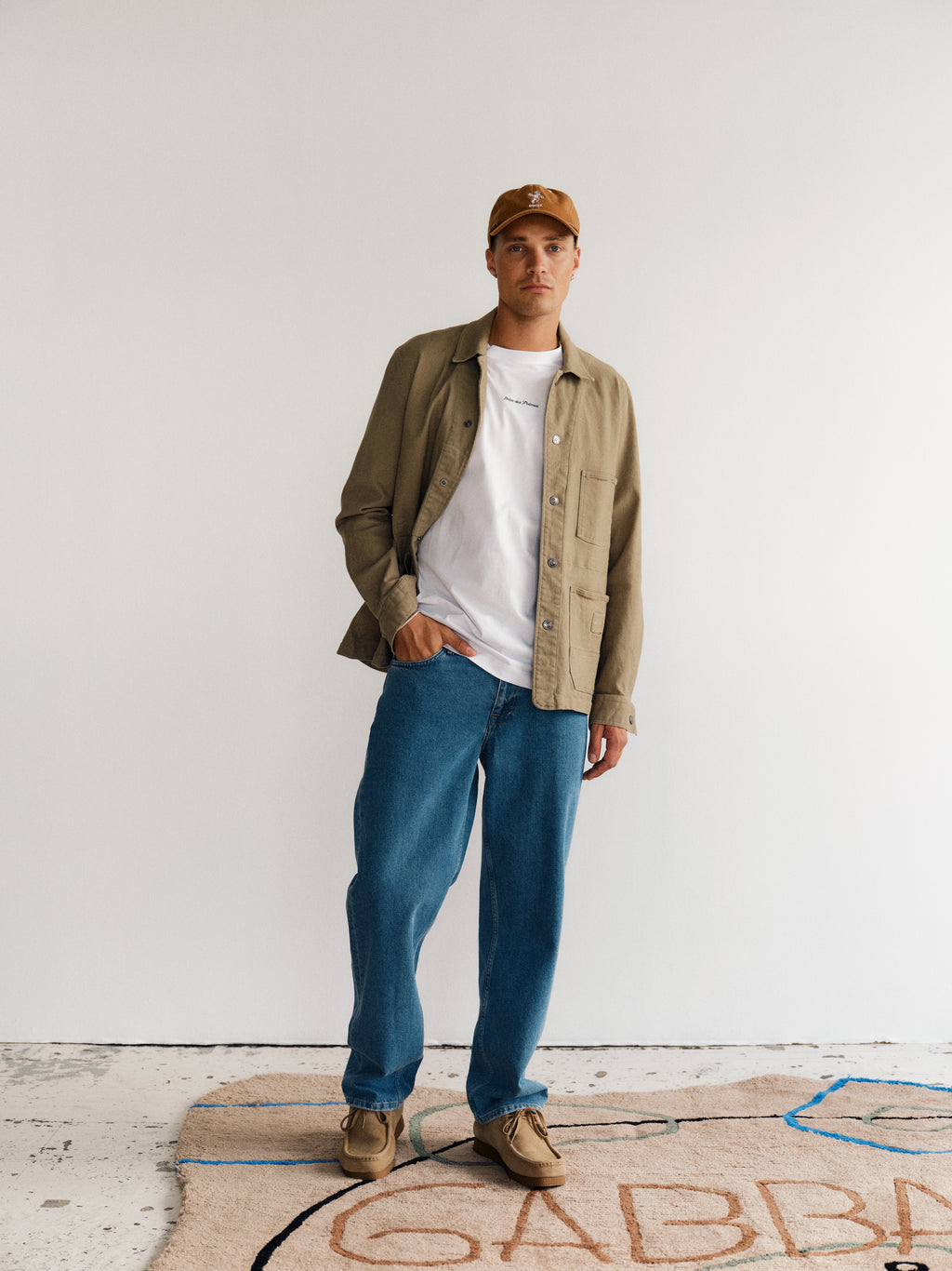 A man stands indoors against a white wall, wearing a tan baseball cap, tan jacket over a white tee, Gabba GABmax 5poc Mid Jeans in Mid Blue Denim, and tan shoes. He stands on a patterned rug with visible lettering.