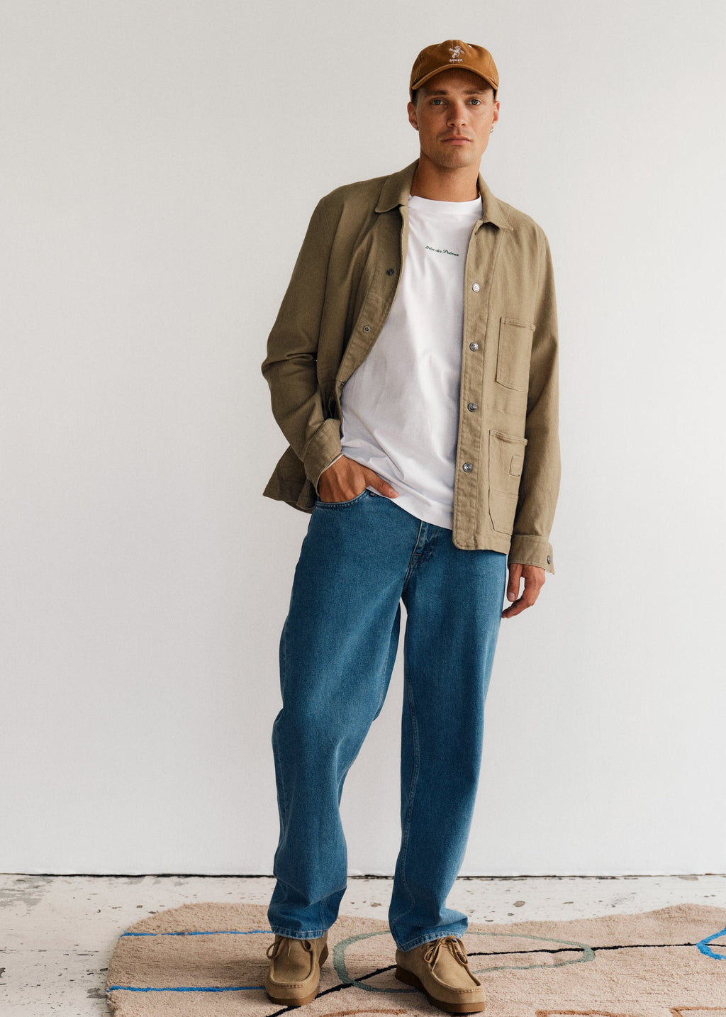 A man stands indoors on a patterned rug, wearing the Gabba GABbeta Twill overshirt in green over a white t-shirt, blue jeans, tan shoes, and a tan baseball cap against a white wall while looking at the camera.