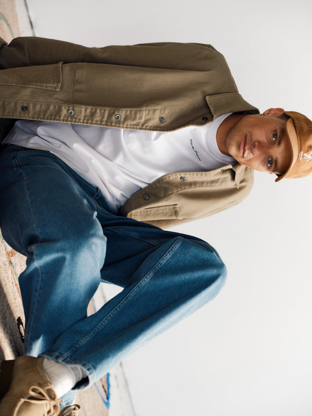 A young man in a tan jacket, blue jeans, tan cap, and a Gabba GABnigel NBH SS T-Shirt GOTS in white sits casually on the floor, looking towards the camera with a relaxed expression.