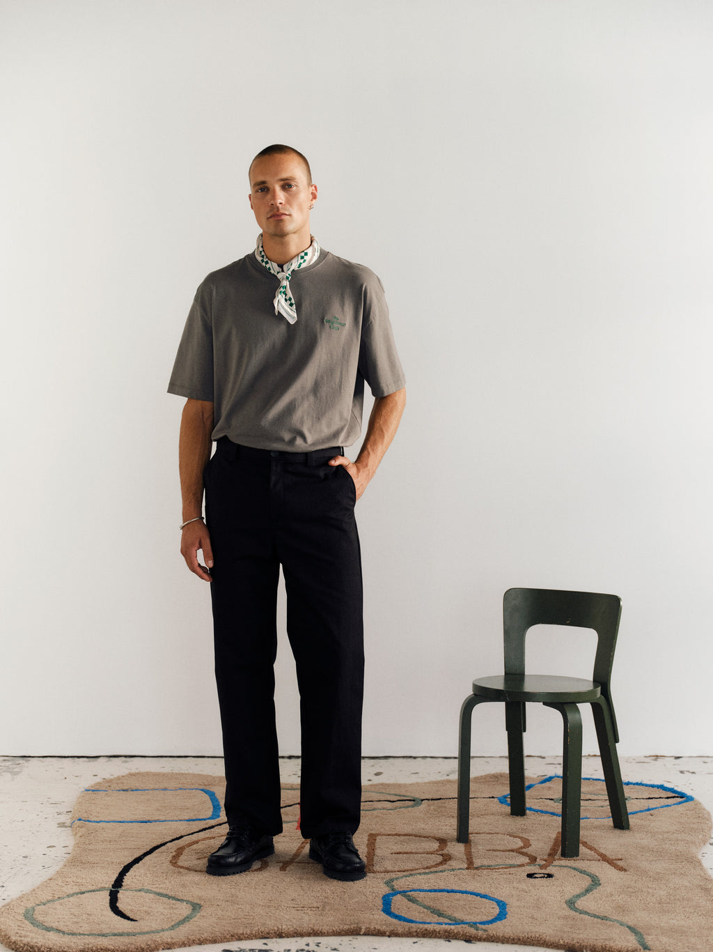 A person stands indoors on a patterned rug, wearing a grey t-shirt, Gabba GABdenver Fuggies Pant in black, black shoes, and a green and white neck scarf. A small green chair is to their right with a plain white background.
