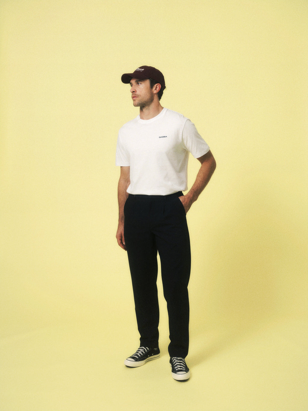 A man wearing a maroon cap, Gabba GABdune Logo SS GOTS t-shirt in white, black pants, and black sneakers stands against a pale yellow background, gazing off to the side with one hand in his pocket.