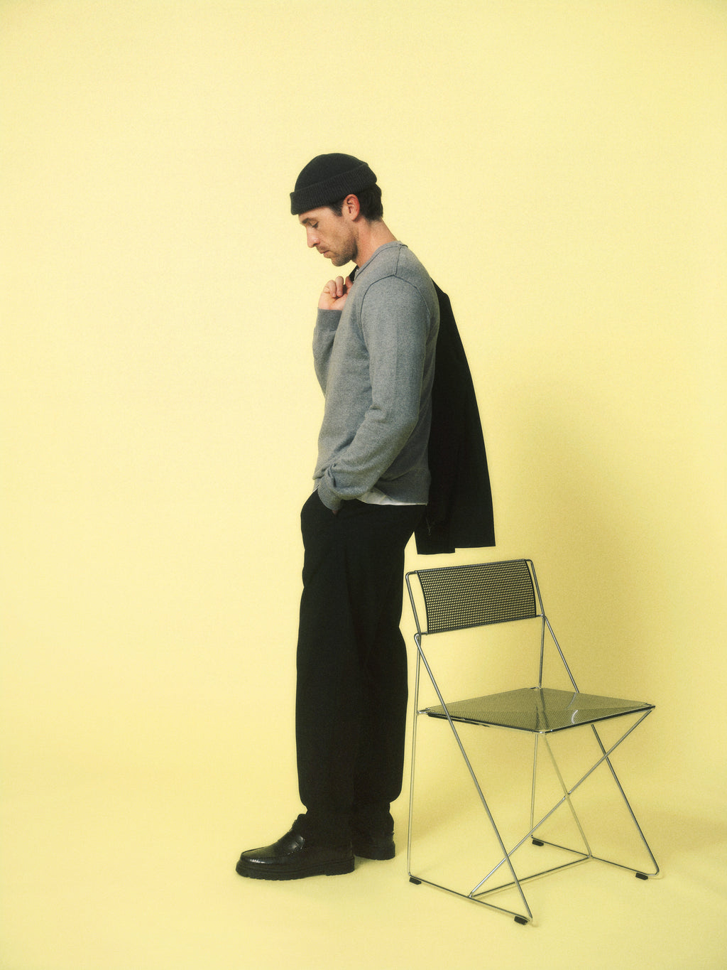 A man wears the Gabba GABnapoli 19 Phin GRS - Black recycled wool sweater with dark pants, black shoes, and a beanie. He stands in profile on a yellow background, holding a jacket next to a folded metal chair.