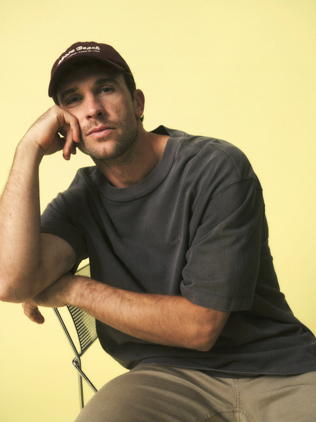 A man in a maroon cap and the Gabba GABnigel Wix Boxy Hour SS tee in dark grey sits on a chair, resting his head on his hand against a pale yellow background, looking at the camera with a relaxed expression.