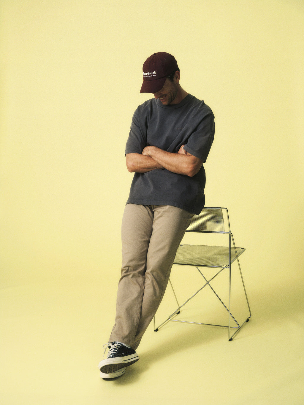 A man in a maroon cap, beige pants, and black sneakers stands before a yellow background, crossing his arms and leaning on a wire chair. He wears the Gabba GABnigel Wix Boxy Hour SS tee in dark grey for a casual modern look.