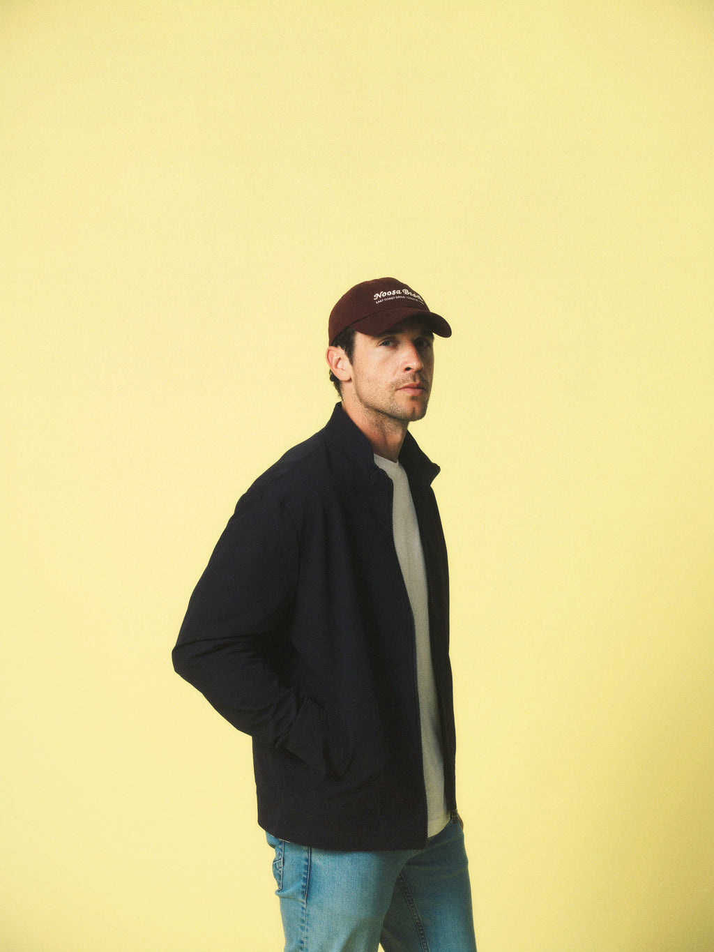 A man wearing the Gabba GABnik Phin GRS - Black jacket, a white shirt, blue jeans, and a brown baseball cap stands against a yellow background, gazing to the side with one hand in his pocket.