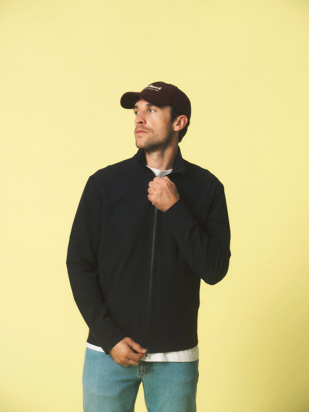 A man in a Gabba GABnik Phin GRS - Black jacket, white shirt, blue jeans, and a dark baseball cap stands against a plain yellow background, looking slightly left while holding his jacket collar.