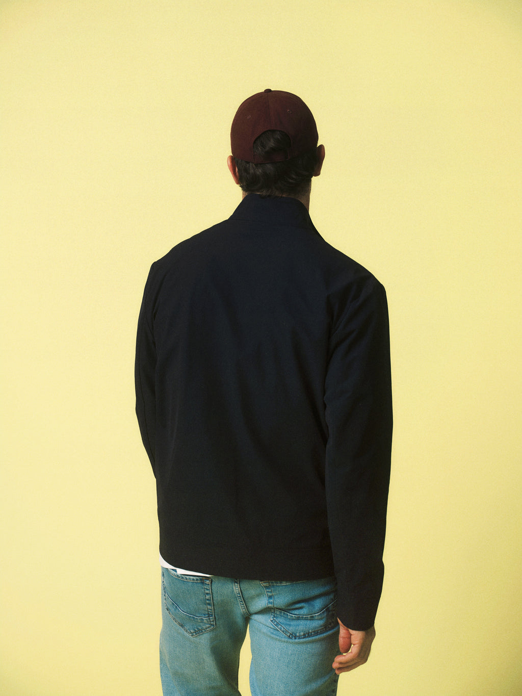 A person wearing the Gabba GABnik Phin GRS Black tailored jacket, a red cap, and blue jeans stands facing away from the camera against a plain yellow background.