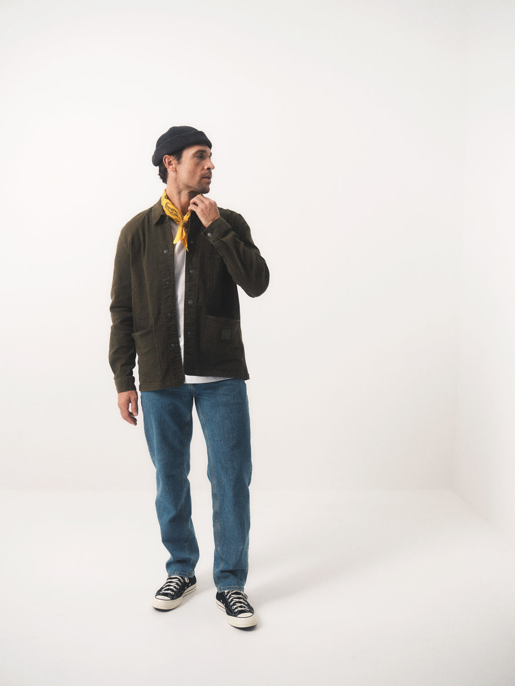 A man stands in a minimalist white room wearing Gabba’s GABmax Cros Lt. Blue Denim jeans, a dark slim-fit jacket, black sneakers, a yellow scarf, and a dark beanie—glancing sideways with one hand touching his scarf.