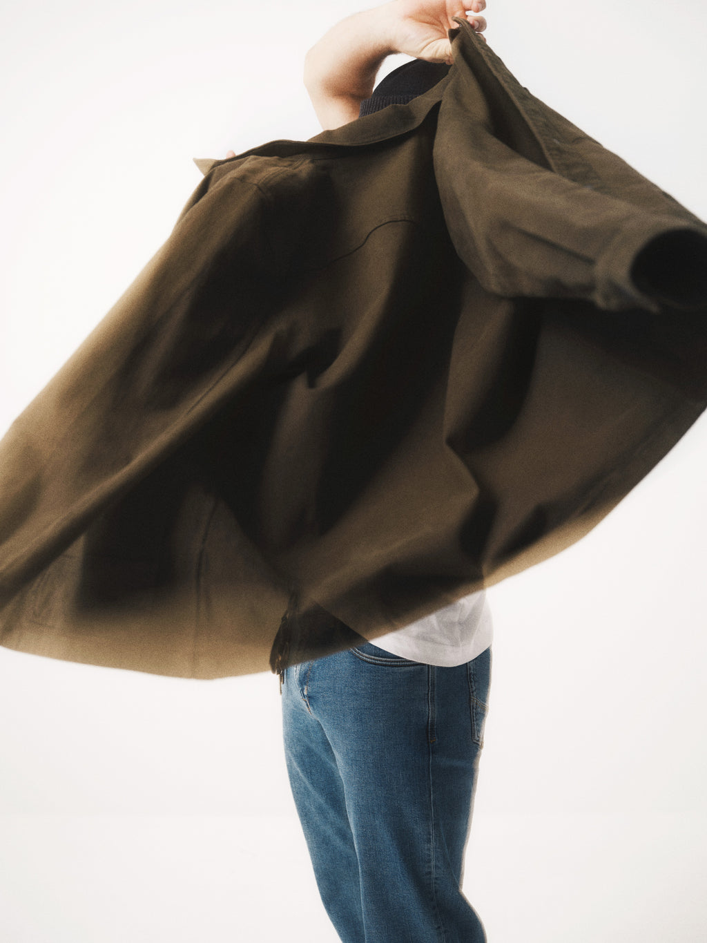 A person in blue jeans and a white shirt is mid-motion putting on or taking off the Gabba GABbeta Twill - Army overshirt, their face mostly obscured by the piece, set against a plain white background.