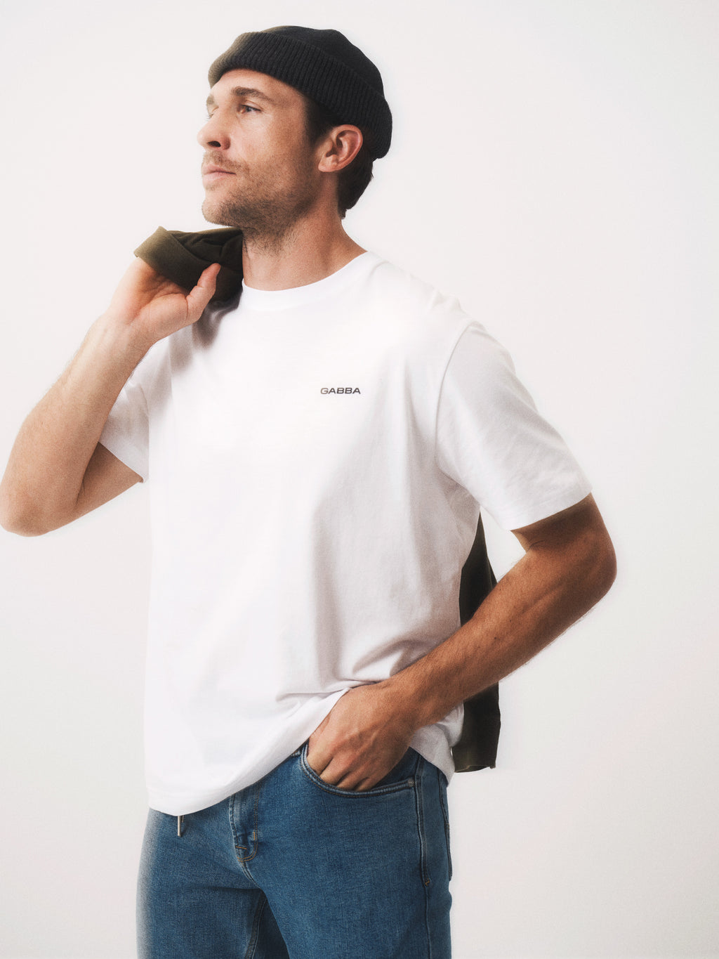 A man in a black beanie, blue jeans, and the Gabba GABdune Logo SS GOTS - White T-shirt stands with one hand in his pocket and a jacket over his shoulder, gazing thoughtfully to the side against a light background.