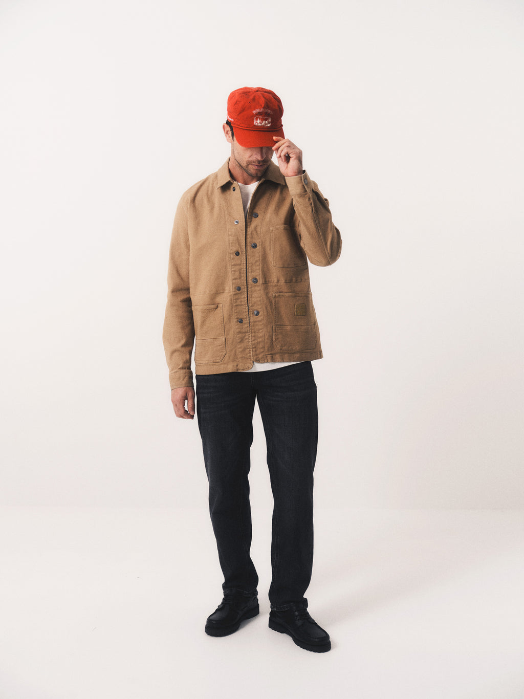 A person stands against a white background wearing the Gabba GABbeta Twill - Sand overshirt, dark jeans, black shoes, and a red cap, adjusting the cap with one hand and tilting their head slightly downward.