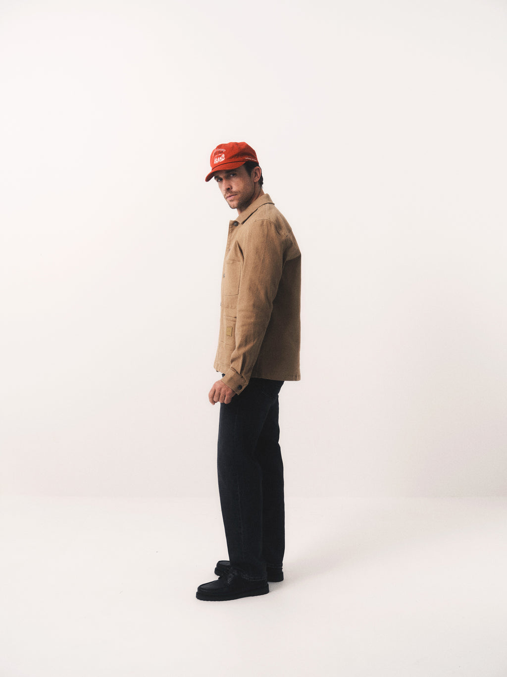A person wearing the Gabba GABbeta Twill jacket in Sand, paired with black pants, black shoes, and a red cap, stands in a minimal white studio space looking over their shoulder at the camera.