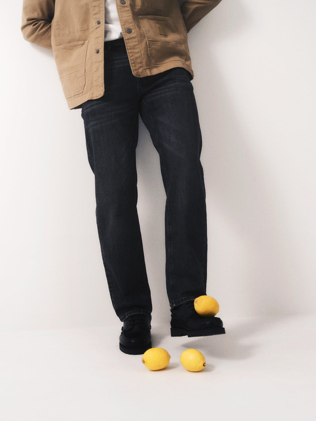 A person, upper body and face not visible, wears a tan jacket, Gabba GABmax F1011 - Black Denim jeans, and black shoes while standing against a white wall. Three lemons are scattered on the floor near their feet.