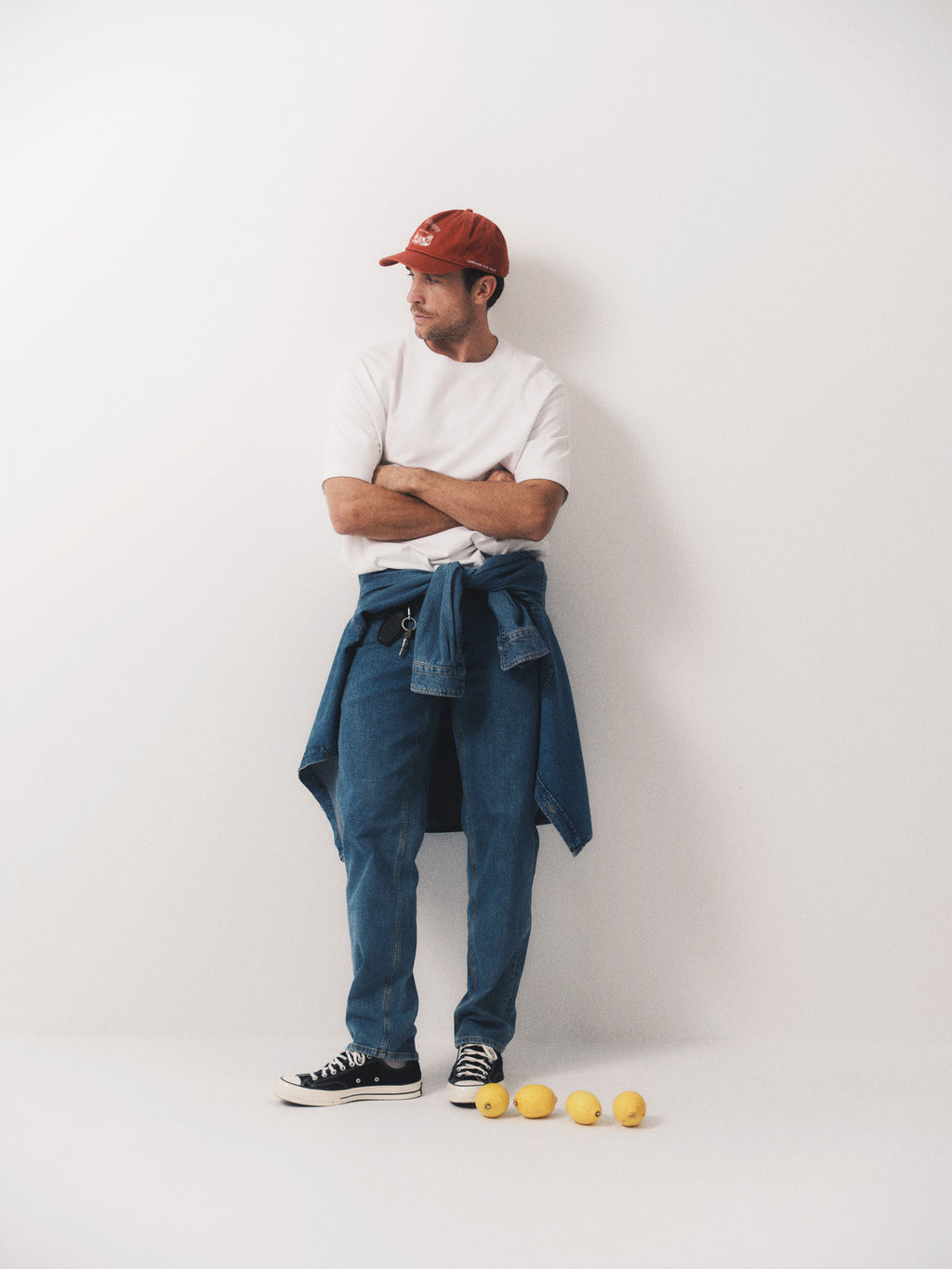 A man in a red cap, white t-shirt, and Gabba GABmax Cros – Lt. Blue Denim jeans stands with arms crossed against a white wall, a denim jacket tied around his waist and four lemons lined up on the floor in front of him.
