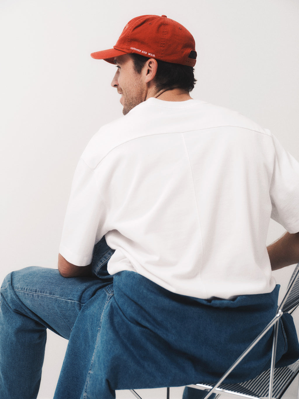 A man in a red cap, Gabba GABnigel Wix Boxy Hour SS white tee, and blue jeans sits casually on a modern metal chair, facing away from the camera against a plain white background.