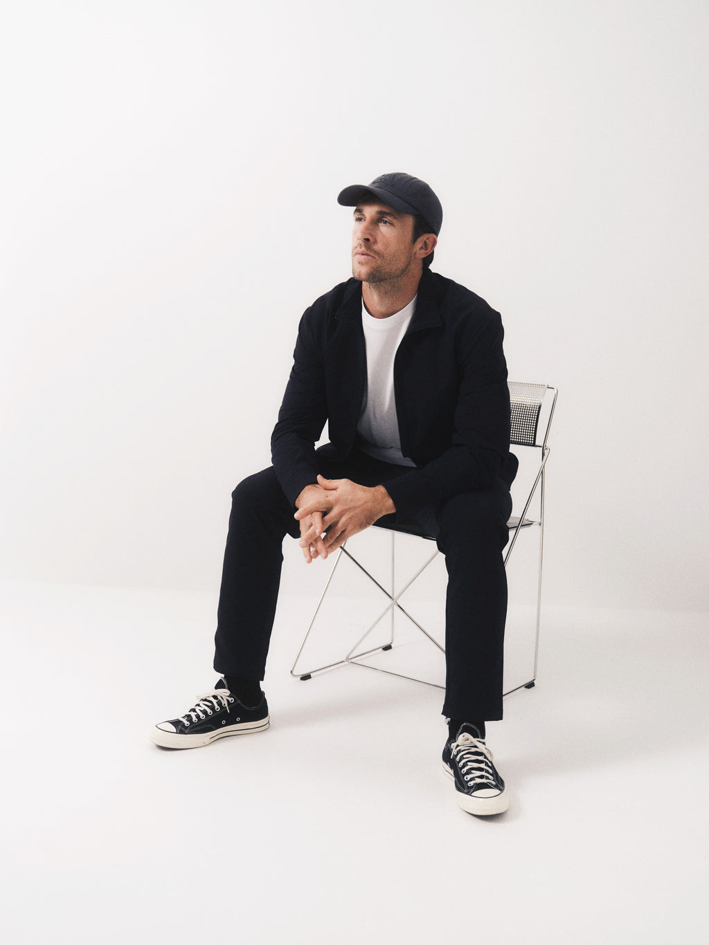 A man in the Gabba GABnik Tech - Black jacket, black cap, white t-shirt, black pants, and black sneakers sits thoughtfully on a folding chair against a plain white background, gazing slightly upward.