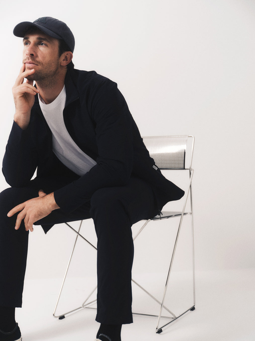 A man wearing the Gabba GABmonza Tech - Black jacket, relaxed fit pants, and a cap sits on a metal chair against a white background, resting his chin on his hand and gazing thoughtfully into the distance.