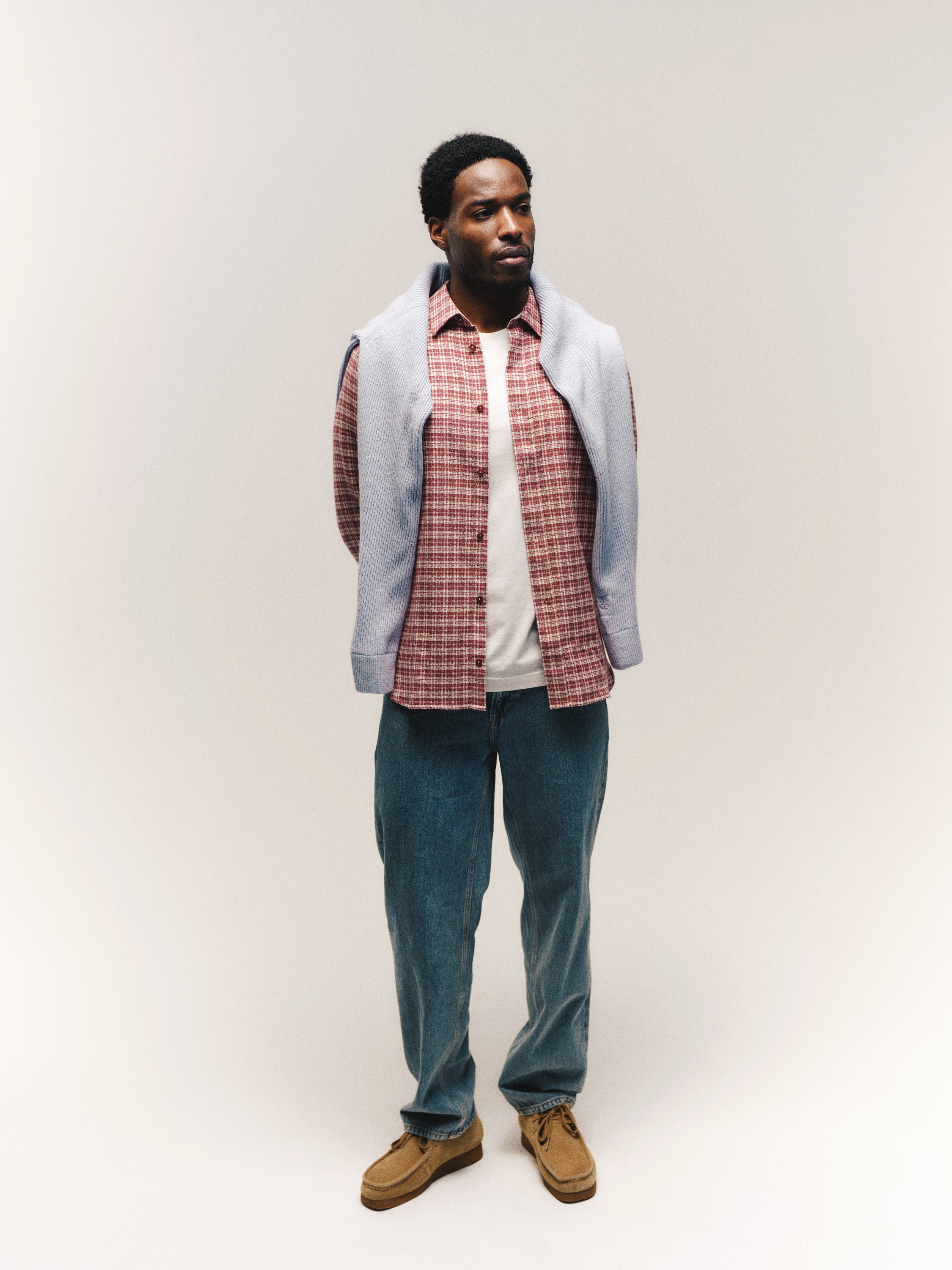 A man stands against a plain background wearing the Gabba GABsven Reg TX2 LS Shirt in Brick Red over a white T-shirt, blue jeans, brown shoes, and a light blue sweater draped over his shoulders.