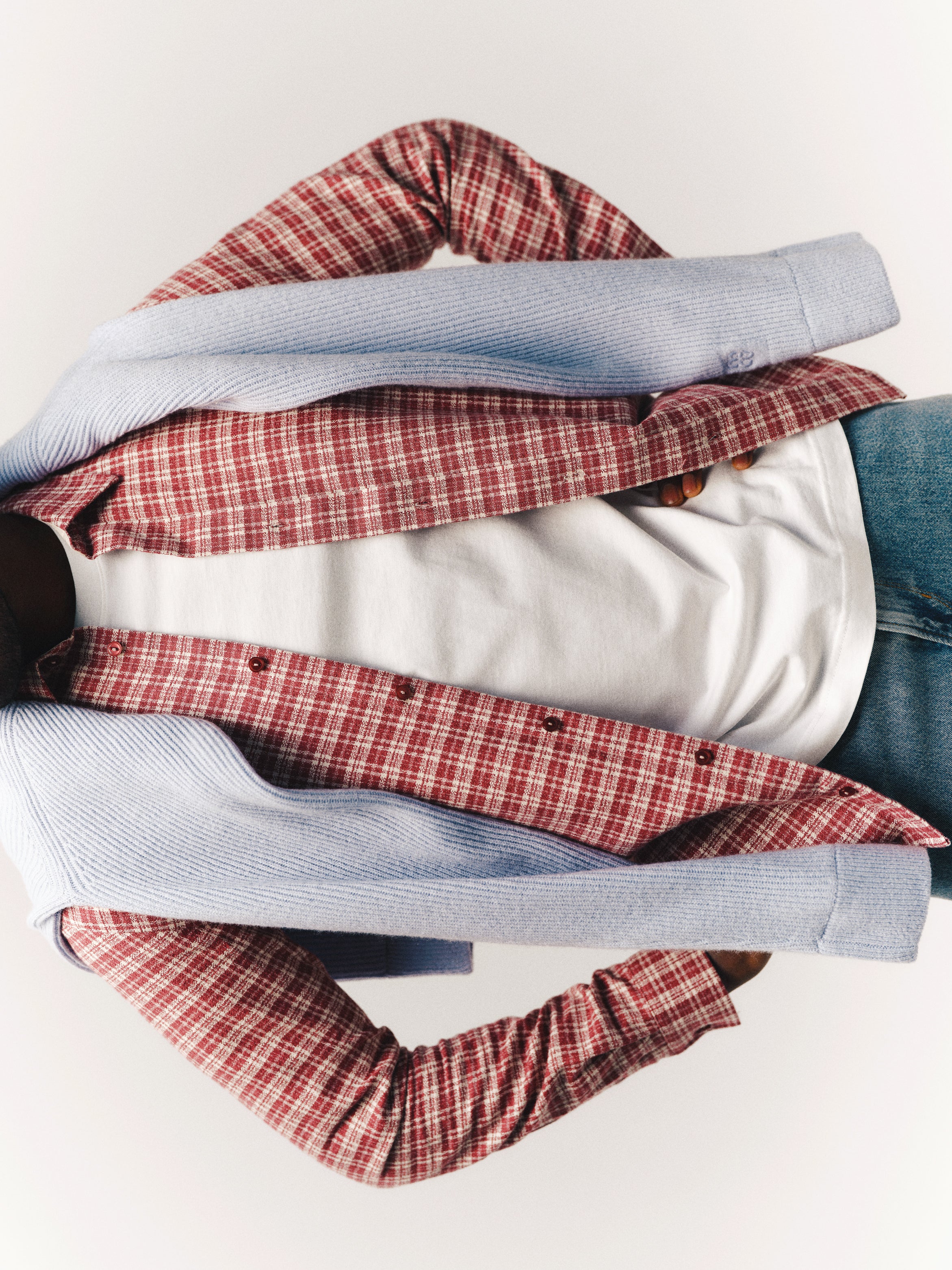 A person wearing the Gabba GABsven Reg TX2 LS Shirt in Brick Red—a regular-fit, yarn-dyed cotton plaid shirt with a checked texture—over a white t-shirt, with a blue sweater draped over their shoulders and paired with blue jeans. The face is not visible in the image.