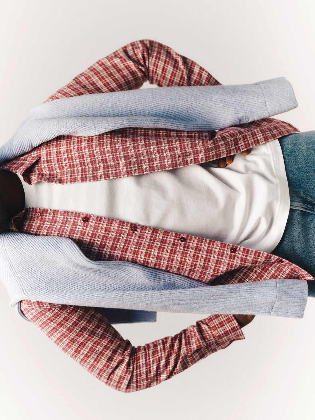A person wearing the Gabba GABsven Reg TX2 LS Shirt in Brick Red—a regular-fit, yarn-dyed cotton plaid shirt with a checked texture—over a white t-shirt, with a blue sweater draped over their shoulders and paired with blue jeans. The face is not visible in the image.
