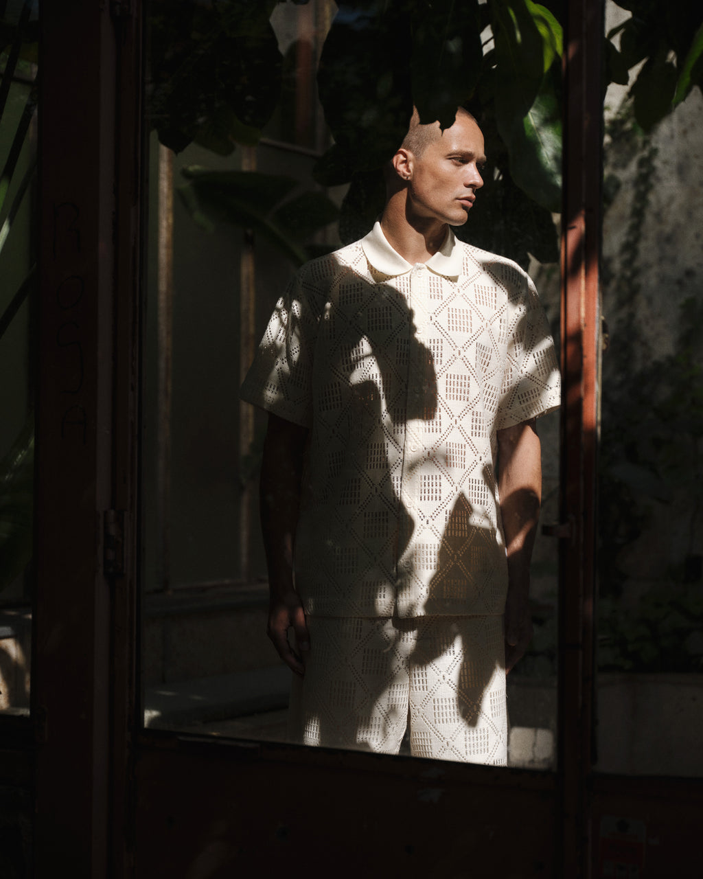 A person in Gabba’s GABjoli Ted Croch Shorts GRS – Birch, paired with a relaxed fit, patterned shirt, stands in dappled sunlight partially shaded by large leaves inside a rustic space with glass doors.