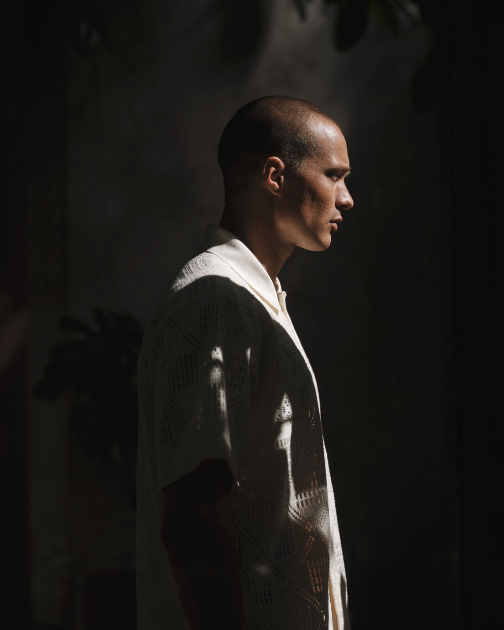A man with a shaved head stands in profile, wearing a white short-sleeve shirt and Gabba GABjoli Ted Croch Shorts GRS in Birch. Sunlight highlights him amid shadows, creating contrast against the dimly lit background.
