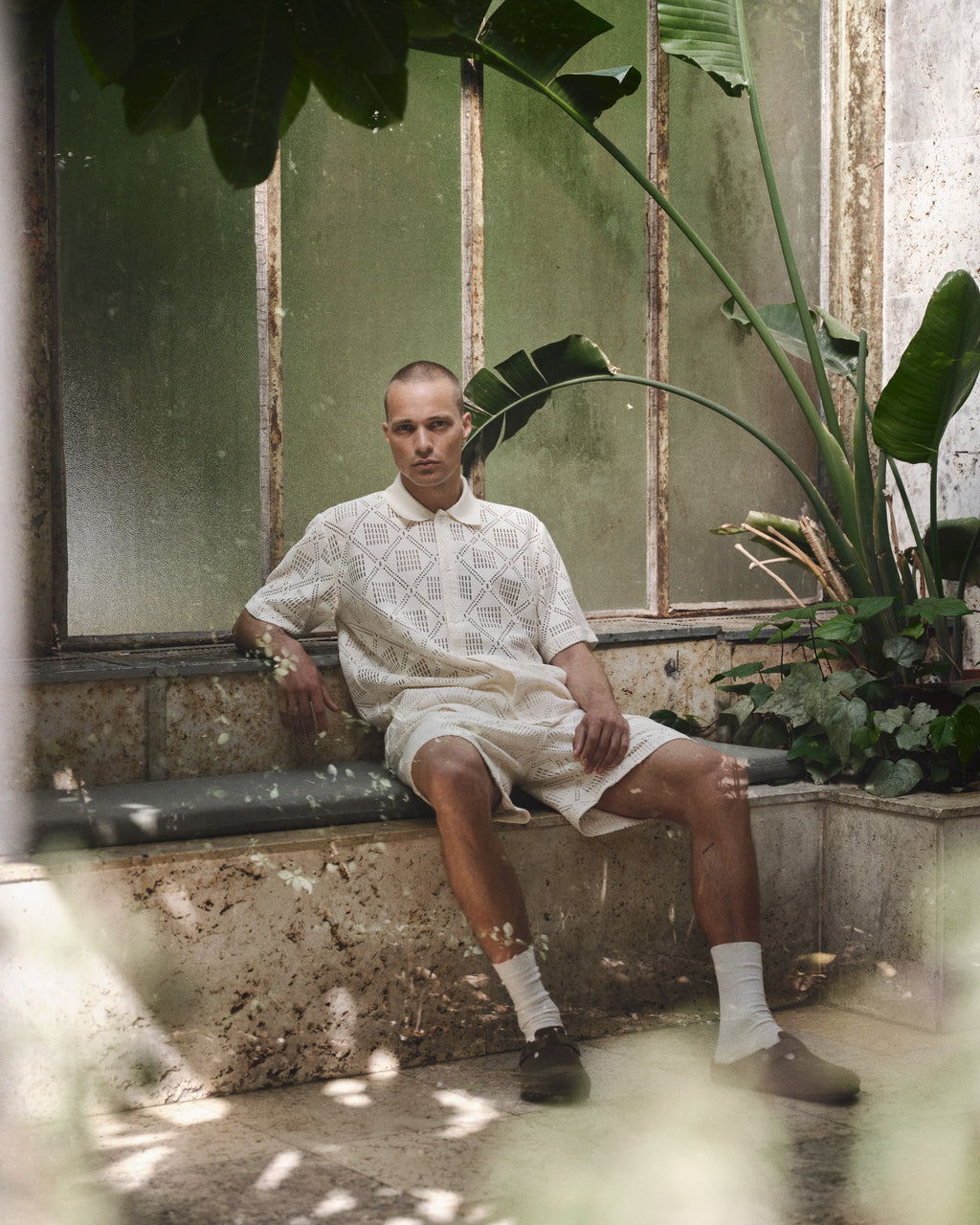 A person with a shaved head sits on a stone bench in a sunlit greenhouse, wearing the Gabba GABsteven Croch SS Shirt GRS - Birch, paired with matching shorts, white socks, and brown shoes amid lush tropical plants.