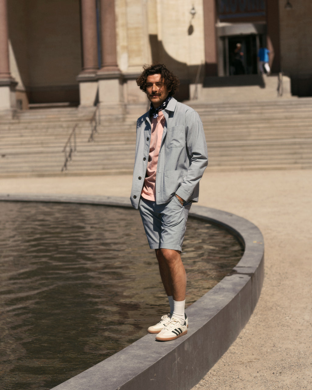 A man with curly hair stands by a round water fountain, wearing Gabba Jet Com2 Shorts in navy, a light gray jacket, pink shirt, and white sneakers. Stone steps and a building facade can be seen in the background.