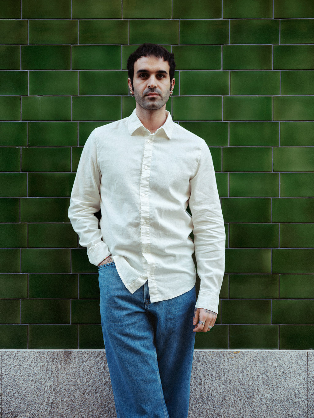 Someone wearing the Gabba GABsven Reg Lins LS Shirt in Off White, paired with blue jeans, stands before a green tiled wall with one hand in their pocket and a neutral expression.