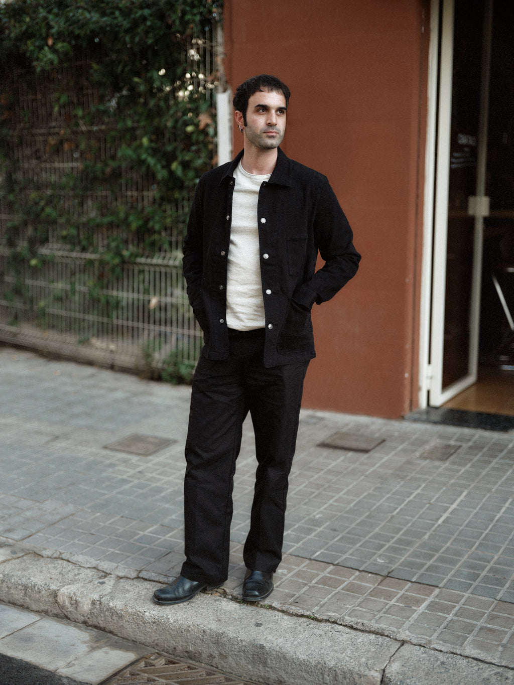 A man stands on a sidewalk near a reddish wall and green plants, wearing the Gabba GABbeta Twill - Black workwear overshirt, a white shirt, and black pants with his hands in his pockets.