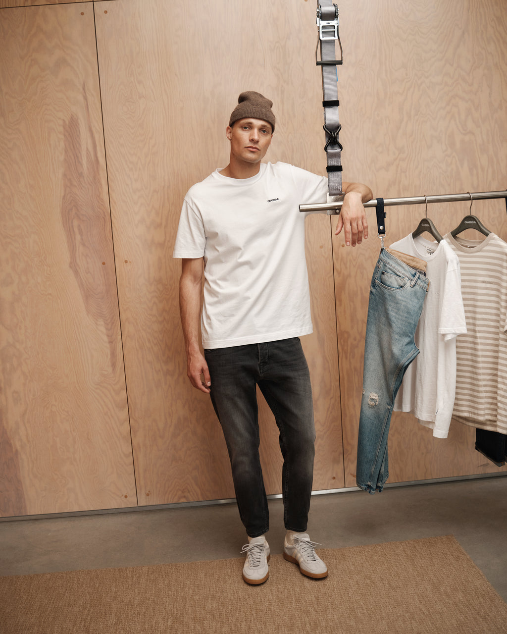 A man in a white T-shirt, GABBA Alex Thor Cropped Jeans - Denim wash, and a gray beanie leans on a clothing rack with casual clothes in a modern, minimalist room with wooden walls.