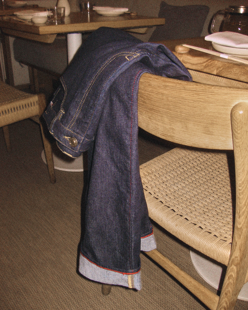 Noah Self - Dark Blue Denim jeans by Gabba, featuring a loose straight fit with rolled cuffs and crafted from sustainable denim, draped over a wooden chair at a dining table set with dishes and glasses.
